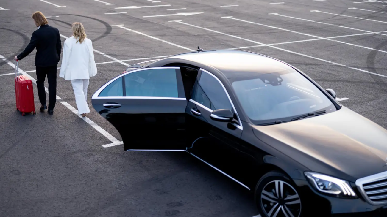 High angle view of a professional man in a black suit rolling a red suitcase alongside a woman in a white pantsuit as they walk away from a black luxury executive sedan with an open rear door in a parking lot