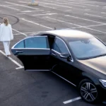 High angle view of a professional man in a black suit rolling a red suitcase alongside a woman in a white pantsuit as they walk away from a black luxury executive sedan with an open rear door in a parking lot