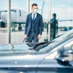 A businessman wearing a blue three piece suit and a medical face mask walks out of an airport terminal toward a waiting luxury car for airport transfer with an airplane visible on the tarmac in the background